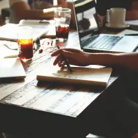 Zoomed in image of hands around a wooden table with notebooks, coffee, and laptops surrounding them.