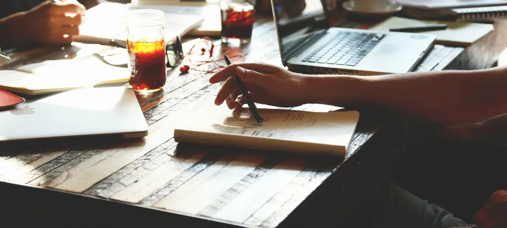 Zoomed in image of hands around a wooden table with notebooks, coffee, and laptops surrounding them.